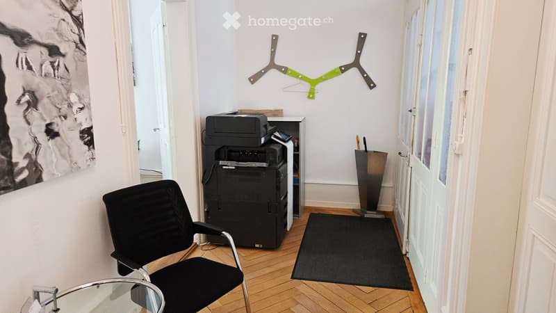 A small, furnished room with a black office chair, a glass table, a printer, and a picture frame on the wall.