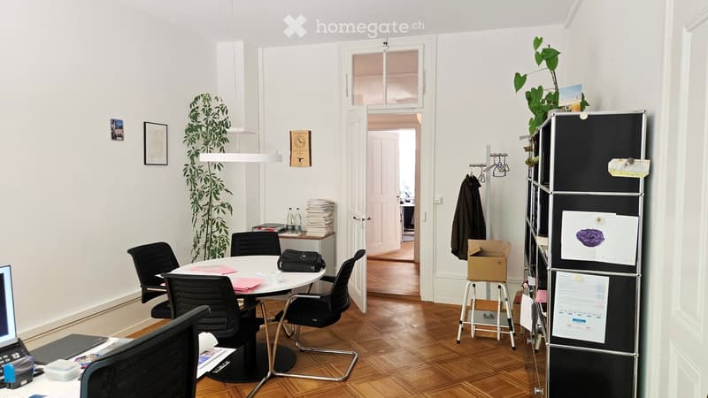 A modern, well-lit office space with a round table surrounded by chairs, a laptop, a refrigerator with papers and drawings, a plant, and a doorway leading to another room.
