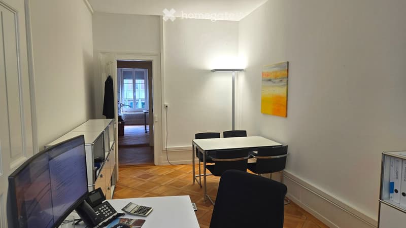 Office space with wooden flooring, white walls, a large screen, telephone, calculator, a table with chairs, a cabinet, and a painting on the wall.