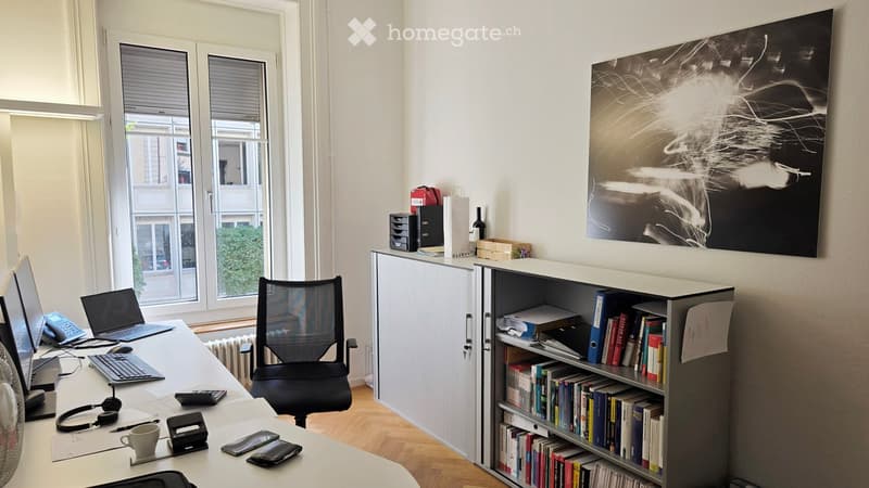 Office space with a desk, ergonomic chair, laptop, keyboard, mouse, headphones, cup, bookshelf filled with books, filing cabinet, and a large abstract picture on the wall. Large windows provide natural light.