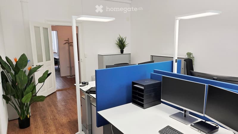 Modern office space with desks, chairs, computer screens, keyboards, and plants. Blue partitions separate workstations.