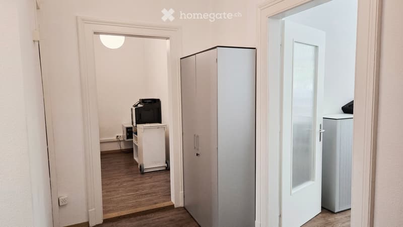 A small, furnished room with a wooden floor, white walls, and a cabinet. A printer sits on a small table, and a refrigerator is visible in the adjacent space.