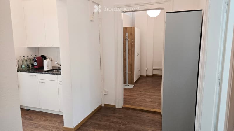 A narrow hallway with wooden flooring leads to a small kitchen area featuring white cabinets, a sink, and various kitchen appliances.