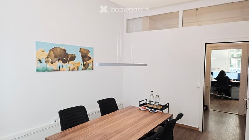 Bright, modern meeting room with a wooden table, black chairs, a large screen, and a picture on the wall. A second room is visible through an open door, containing a desk and computer setup.