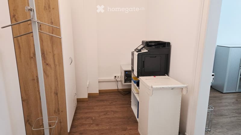 A small, furnished room with a printer on a table, a wooden cabinet, and a shelf with various items.