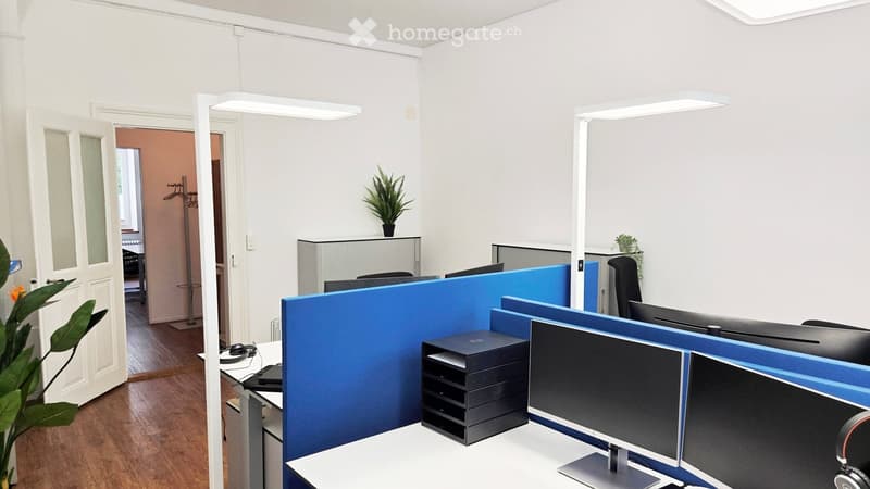 Modern office space with desks, chairs, multiple screens, laptops, and plants.