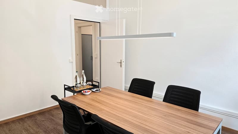 A modern, minimalist room with a wooden table, four black chairs, a small side table with bottles and a bowl, and a sleek light fixture hanging from the ceiling.