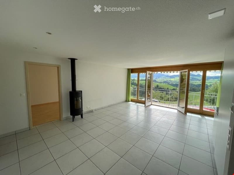 Empty room with tiled floor, white walls, a wood-burning stove, and large windows offering a scenic view of the landscape.