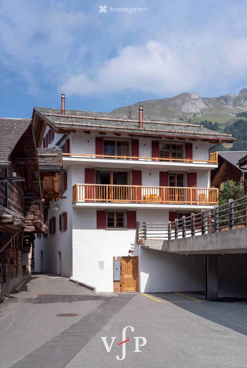 A two-story white building with red shutters and wooden balconies, situated in a mountainous area with a clear blue sky and distant mountains.