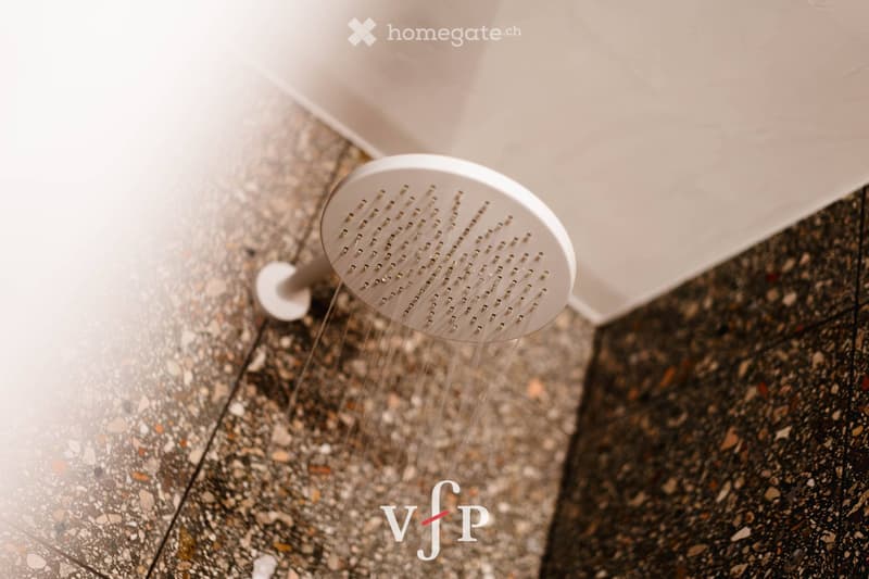 Close-up of a modern white showerhead with multiple spray holes, mounted on a dark, speckled tile wall.