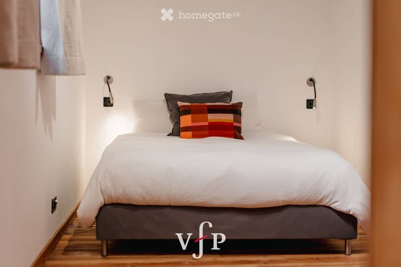 A modern, minimalist bedroom with a neatly made bed, white bedding, and a colorful throw pillow. The room features wooden flooring and wall-mounted lamps on either side of the bed.