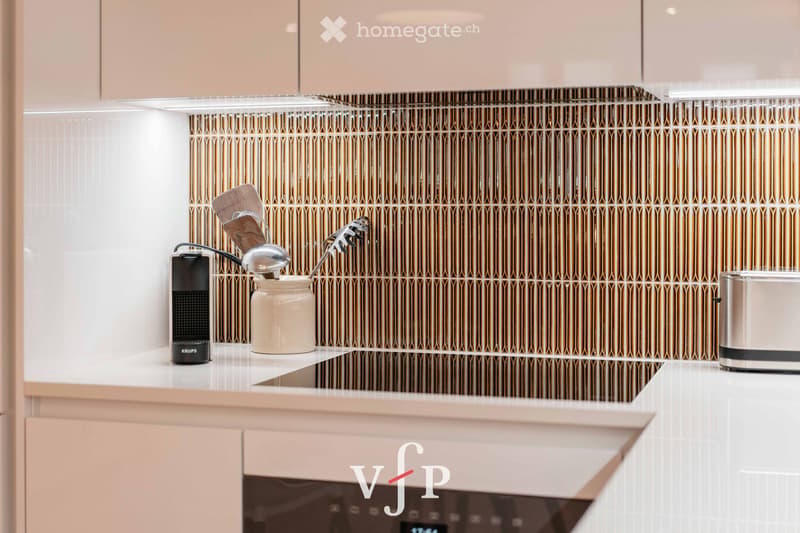Modern kitchen with light-colored cabinets, white countertops, and a decorative backsplash featuring vertical metallic strips. A small speaker, a jar with kitchen utensils, and a toaster are visible on the counter.