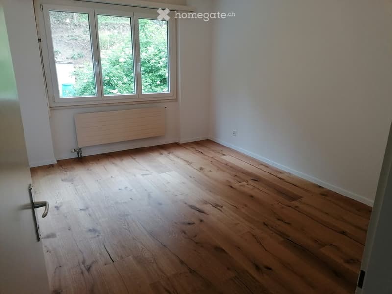 Empty room with light wooden flooring, white walls, and a large window with a view of greenery. A radiator is mounted on the wall below the window.