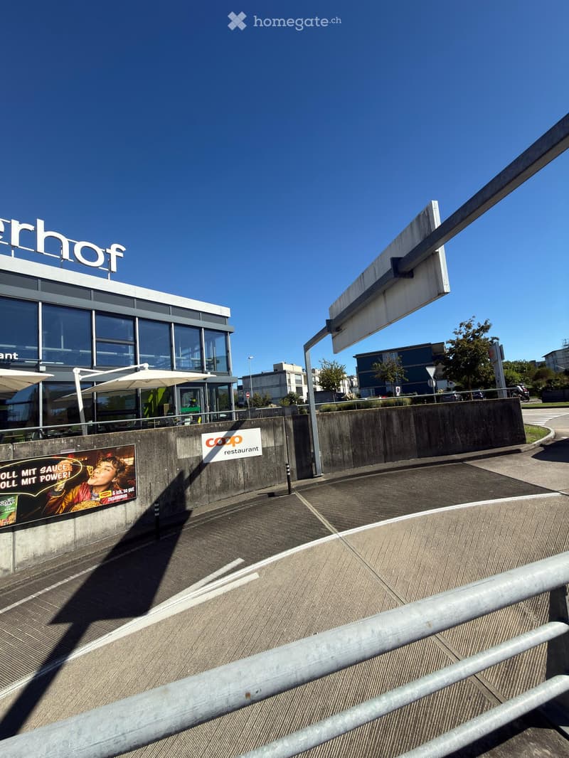 Modern building with large glass windows and a sign reading "Erhof" on the roof. The entrance features outdoor seating with umbrellas and a concrete barrier with advertisements.