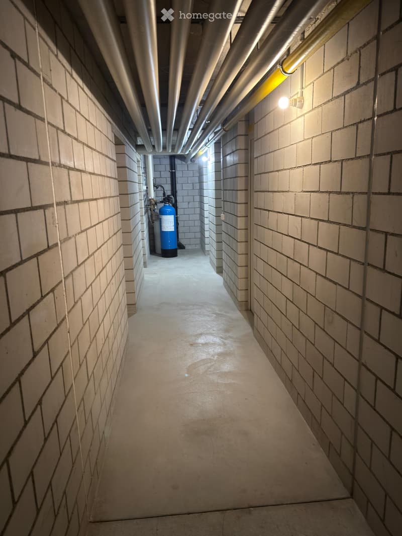 A narrow, industrial-style corridor with brick walls, concrete floor, exposed pipes, and a blue water tank in the background.
