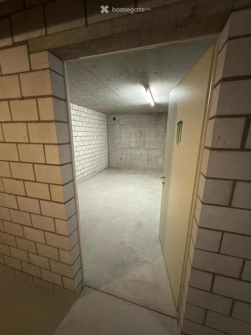 Empty underground parking space with concrete walls, floor, and ceiling, featuring a small window and a single light fixture.