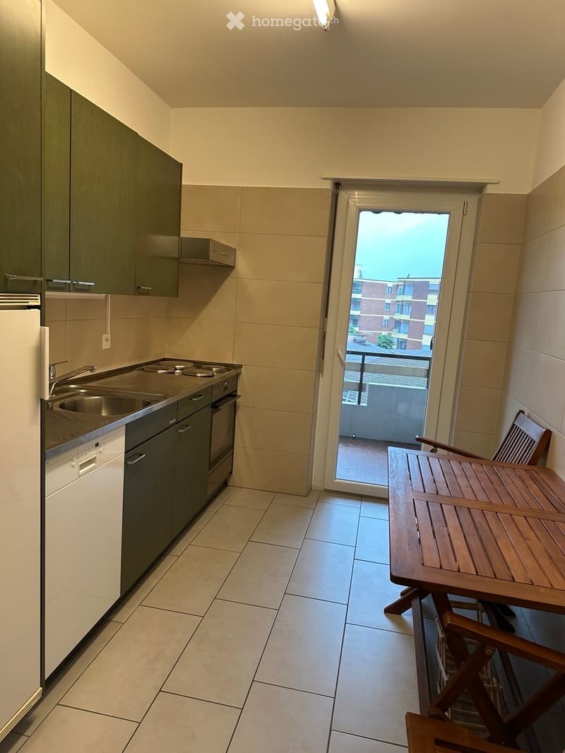 Modern kitchen with stainless steel appliances, a small wooden table and chairs, and a sliding door leading to a balcony.