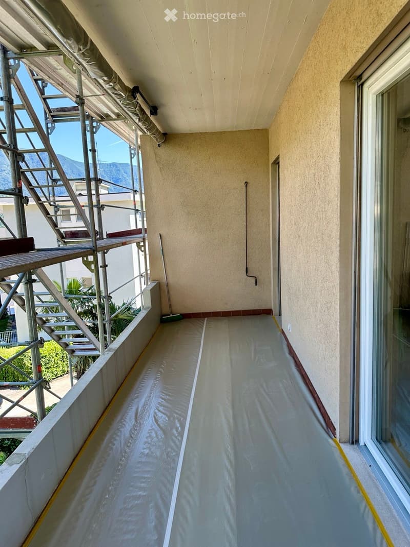 Balcony with a smooth, light-colored floor, beige walls, and a metal staircase on the left side. The balcony is partially covered with protective plastic sheeting and has a view of greenery and mountains in the background.