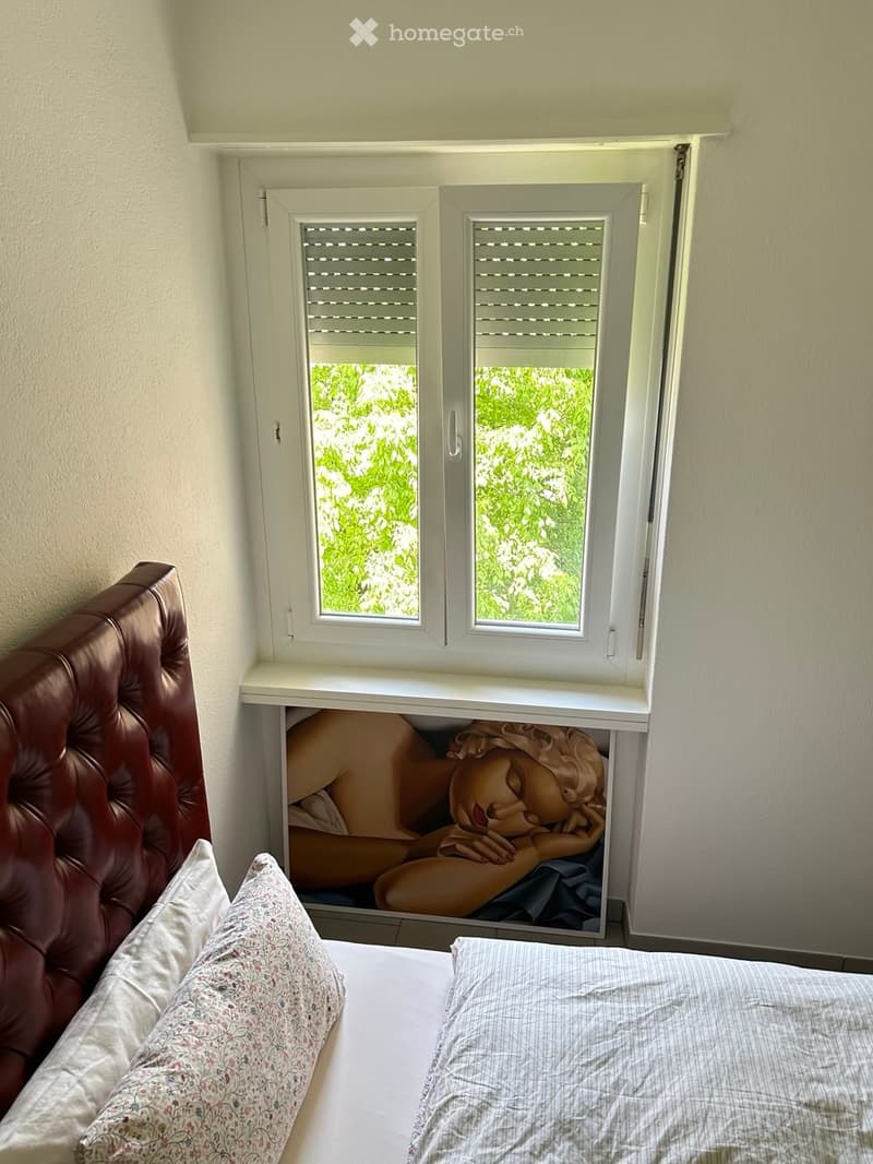 Bedroom with a leather headboard, two pillows, and a framed artwork of a reclining figure on a shelf beneath a window with shutters.