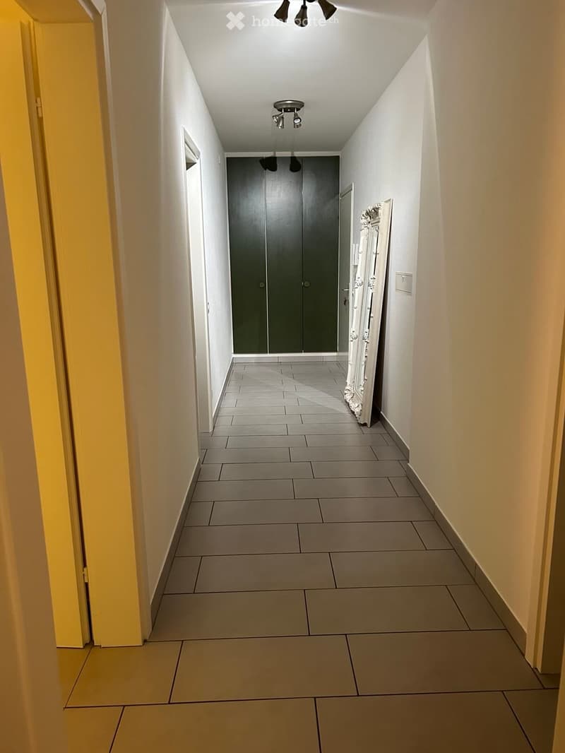 A long, narrow hallway with tiled flooring, white walls, and recessed lighting. Features a large dark green closet door and a decorative mirror on the right side.