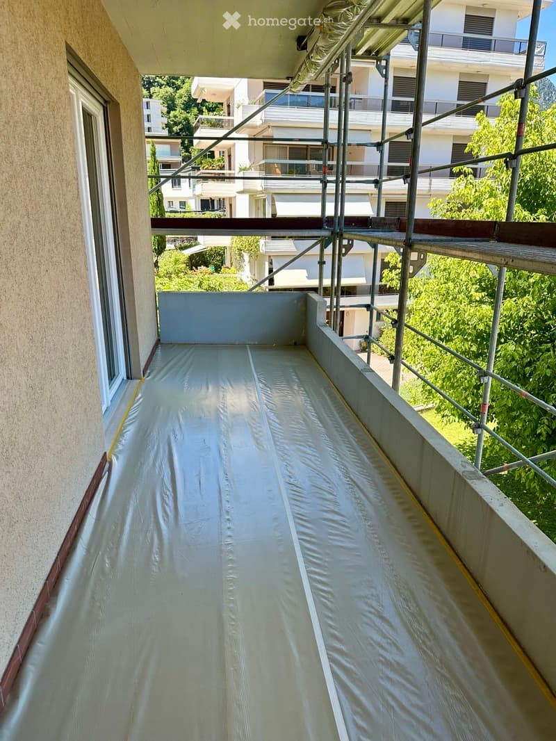 Balcony under construction with scaffolding and protective covering, overlooking a residential area with greenery and neighboring buildings.