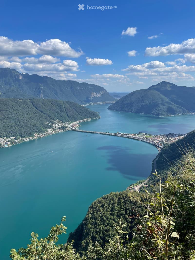 Panoramic view of a lake surrounded by mountains, with a bridge and small town visible along the shoreline.