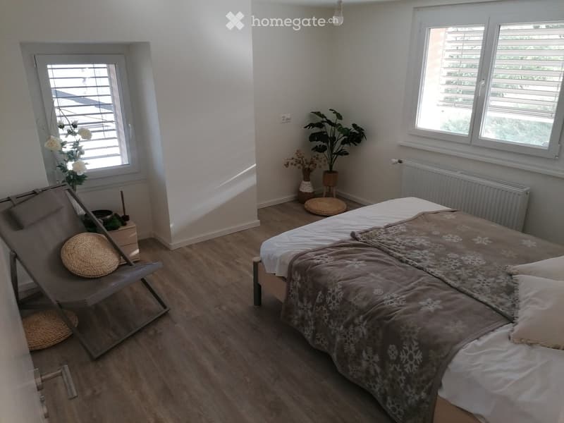 Cozy bedroom with wooden flooring, a double bed with a patterned duvet, a wicker chair, a small nightstand, and a potted plant near the window.