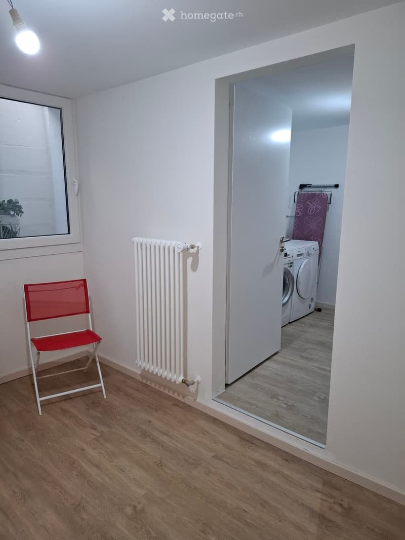 A small, well-lit room with wooden flooring, a white radiator, a red folding chair, and an open doorway leading to a laundry area with a washing machine and dryer.