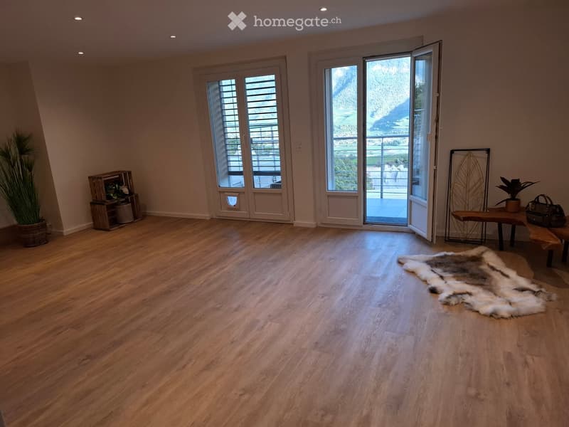 Bright, open room with wooden flooring, large windows, and a balcony view. Includes a bench, a small table with a handbag, a furry rug, a shelf with plants, and a decorative metal wall piece.