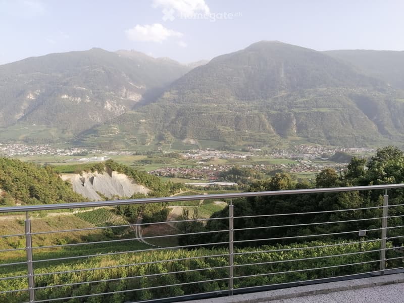 Balcony with metal railing overlooking a scenic valley, mountains, and a town below.