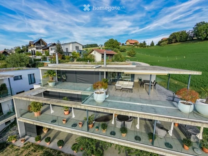 Modern two-story house with a large terrace and glass railings, surrounded by greenery and potted plants. The house features a flat roof and contemporary design.