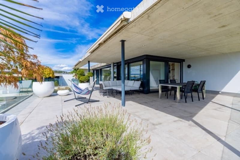 Modern outdoor patio with a covered seating area, white furniture, and a dining set, surrounded by plants and a scenic view.
