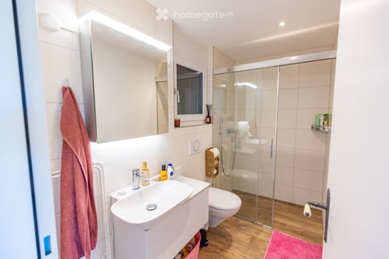 Modern bathroom with a white sink, chrome faucet, toilet, large mirror, and a glass-enclosed shower. The room features light-colored tiles and a wooden floor.