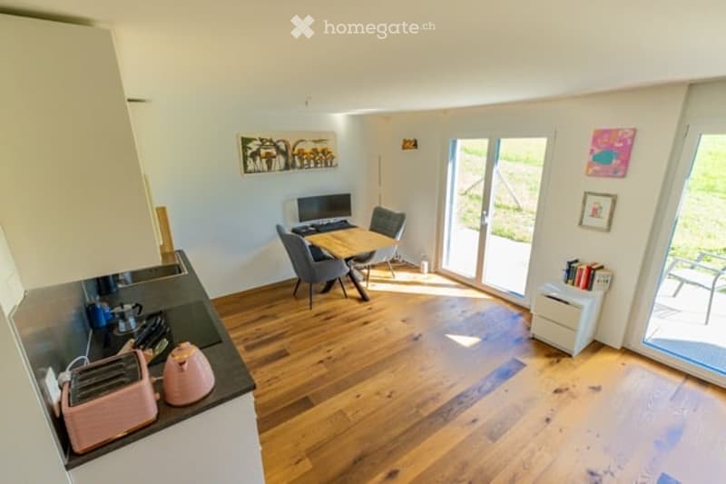 Open-plan kitchen and dining area with light wood flooring, white cabinets, and modern appliances. A small dining table with chairs is positioned near large glass doors leading to a balcony.