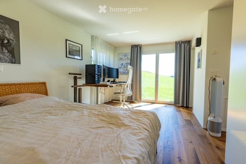 Well-lit bedroom with a large bed, a modern desk setup with a computer and dual monitors, a comfortable chair, and large windows with curtains. The room features light-colored wooden flooring and walls adorned with framed pictures.