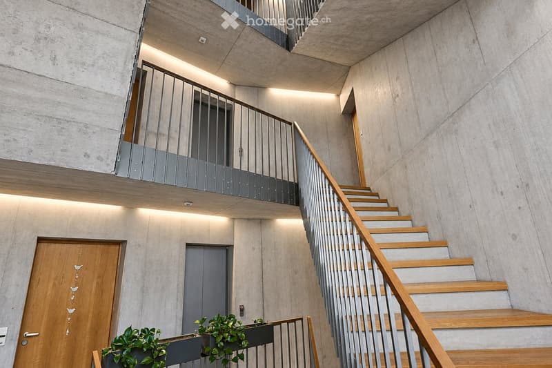 Modern interior staircase with wooden steps, metal railing, and concrete walls. Features recessed lighting and a small balcony area above.