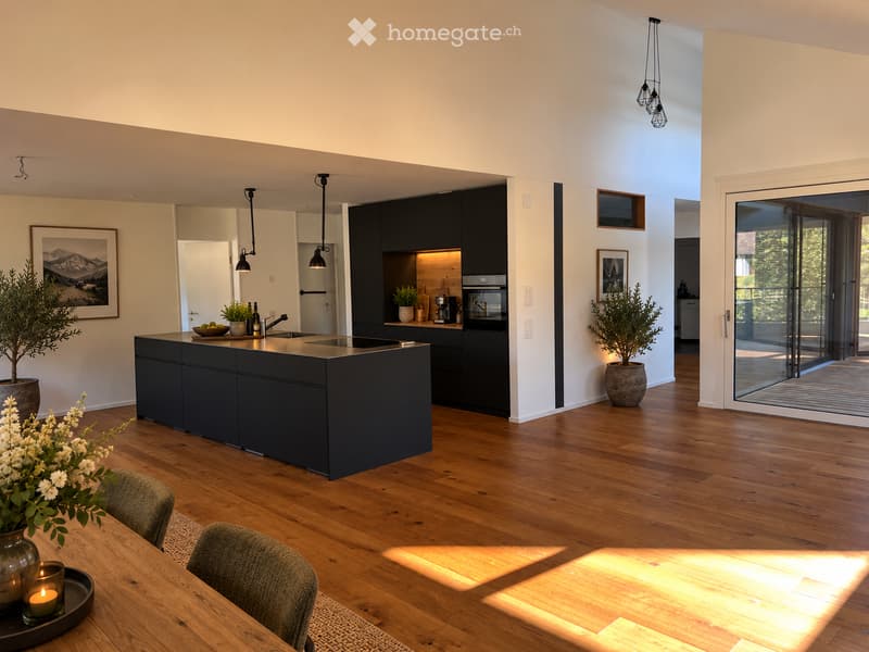 Modern kitchen with dark grey cabinets, a central island, and a dining area with a wooden table and chairs. Features pendant lights, a coffee maker, and potted plants.