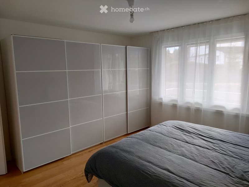 Bedroom with a large grey wardrobe and a neatly made bed with grey bedding, wooden flooring, and large windows with sheer curtains.