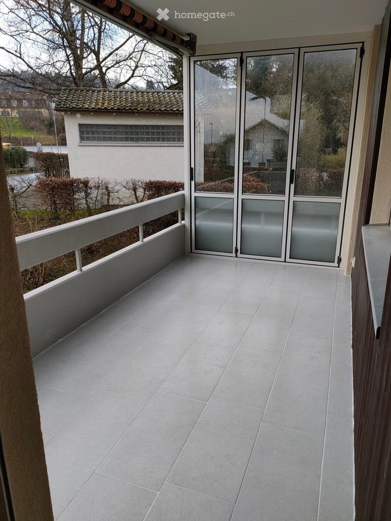 Balcony with tiled floor, glass sliding doors, white railing, and view of neighboring house and garden.