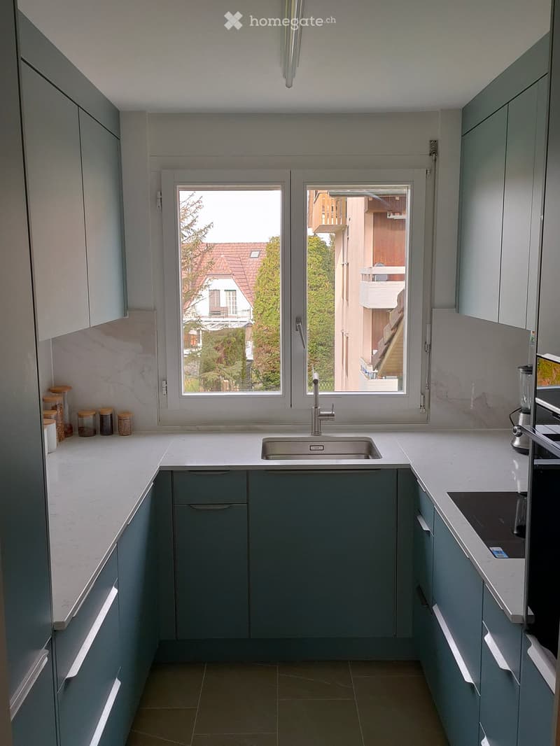 Modern kitchen with grey cabinets, white countertops, and a stainless steel sink. Features a window with a view of a balcony and outdoor area.