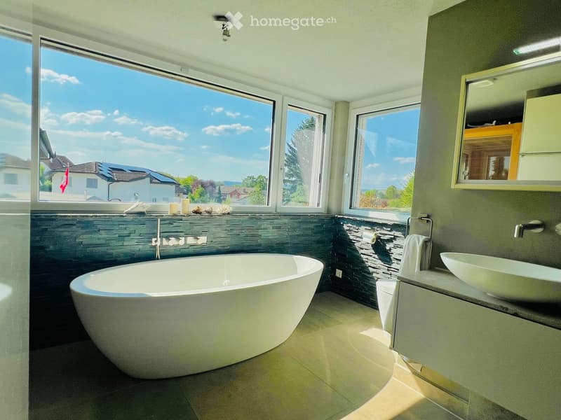 Modern bathroom with a large bathtub, sink, mirror, and towel, featuring large windows with a scenic view.