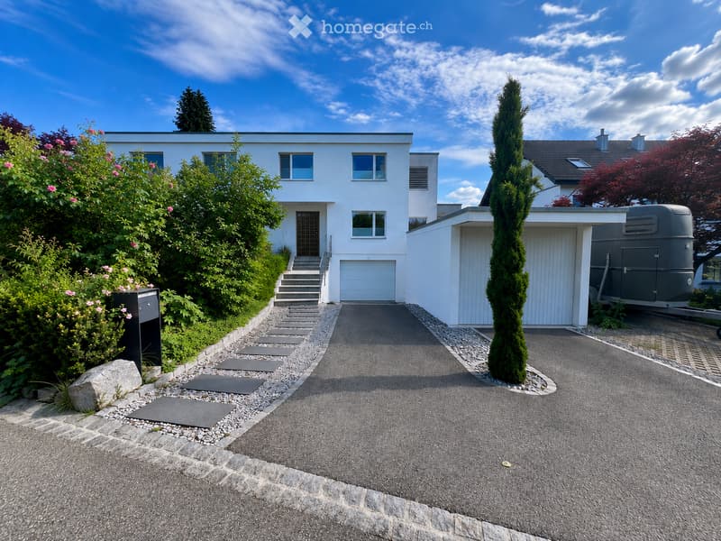 Modern two-story house with white exterior, multiple windows, garage, and landscaped garden with trees and shrubs.