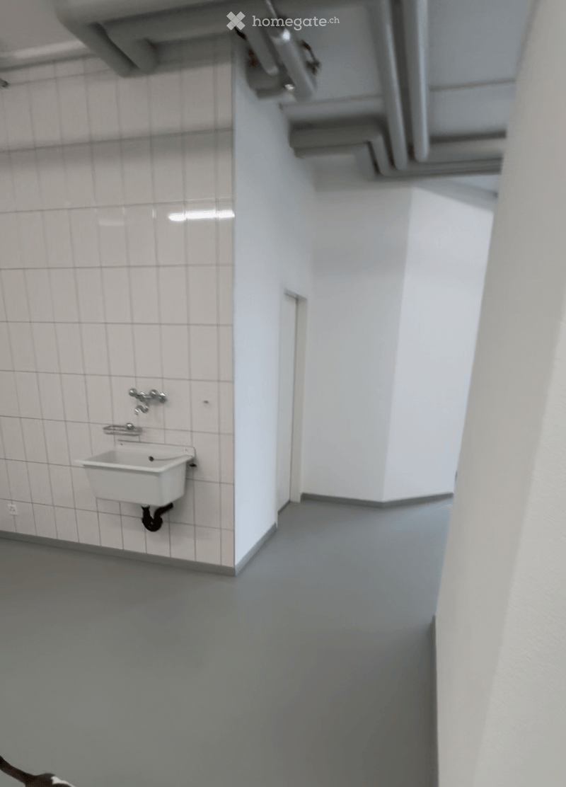 A small, clean bathroom with white tiled walls, a white sink, and a modern faucet. The floor is grey and the space is well-lit.