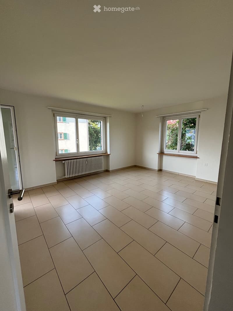 Empty room with two windows, beige tiles, white walls, windows with white frames and green shutters, two radiators below windows.