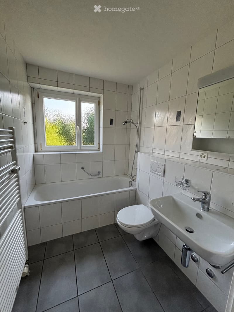 White tiled walls, white bathtub, white toilet, white sink, mirror, towel rack, window, showerhead, power outlet