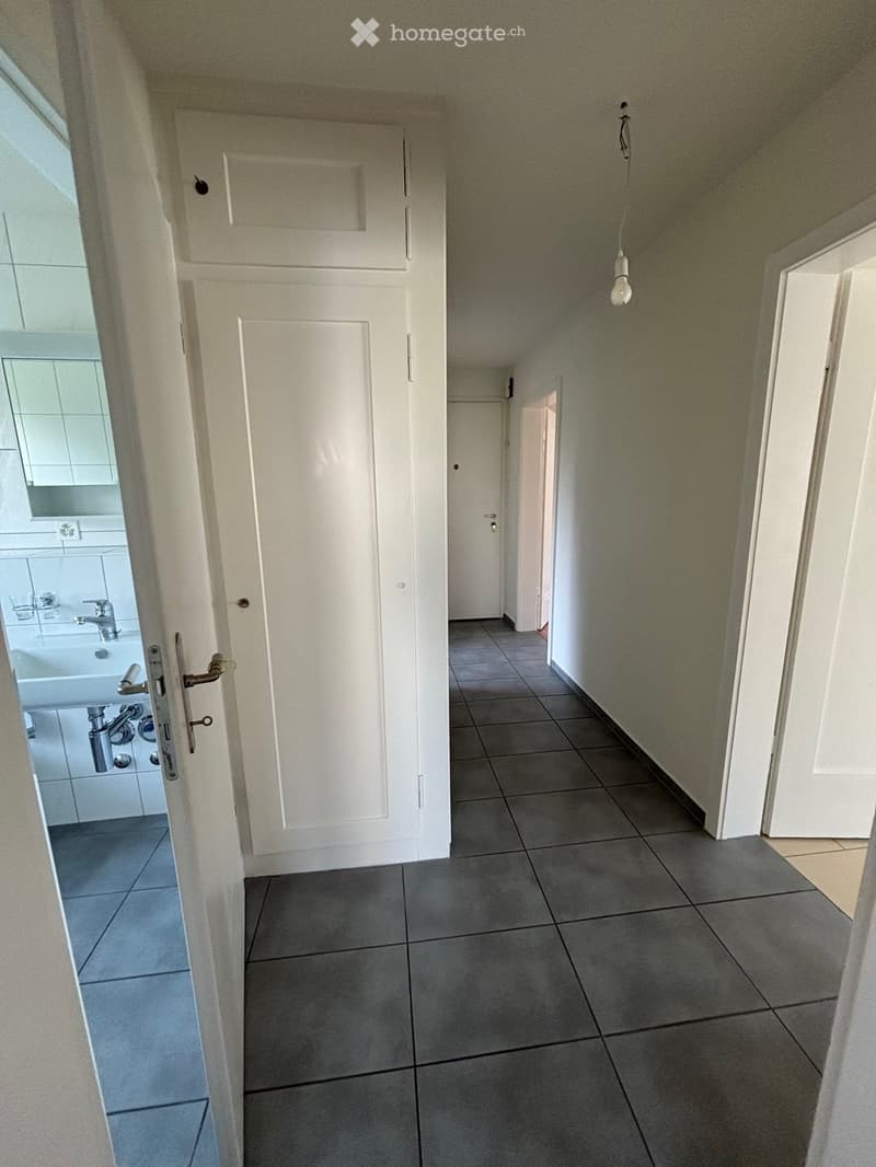 White walls, tiled floor, sink, mirror, dark grey tiles, and an open door leading to another room.