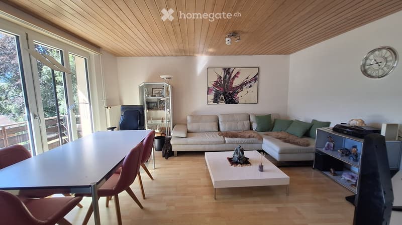 Bright living room with wooden floors, white walls, and large windows. Features a cream-colored sofa with green cushions, a coffee table with a decorative item, a dining table with chairs, a floor lamp, and a picture frame on the wall.