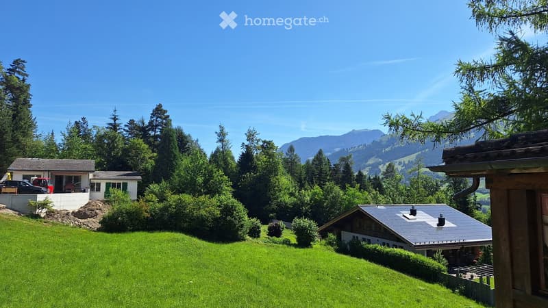 A picturesque house with a grey roof and white walls, situated on a lush green lawn surrounded by trees and mountains in the background.