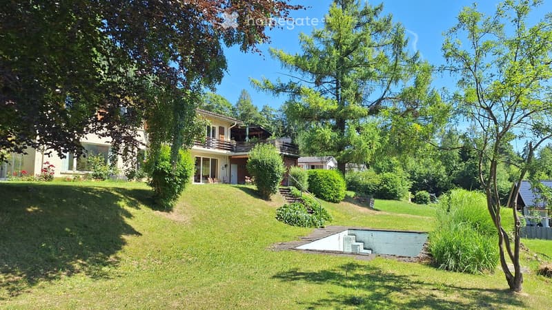 A well-maintained garden with a raised lawn, trees, shrubs, and a small pool area. A two-story house with a balcony and patio is visible in the background.