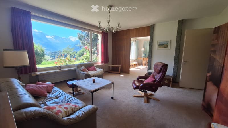 Bright living room with large windows offering a scenic mountain view, furnished with a sofa, coffee table, armchair, and a small table with a lamp.